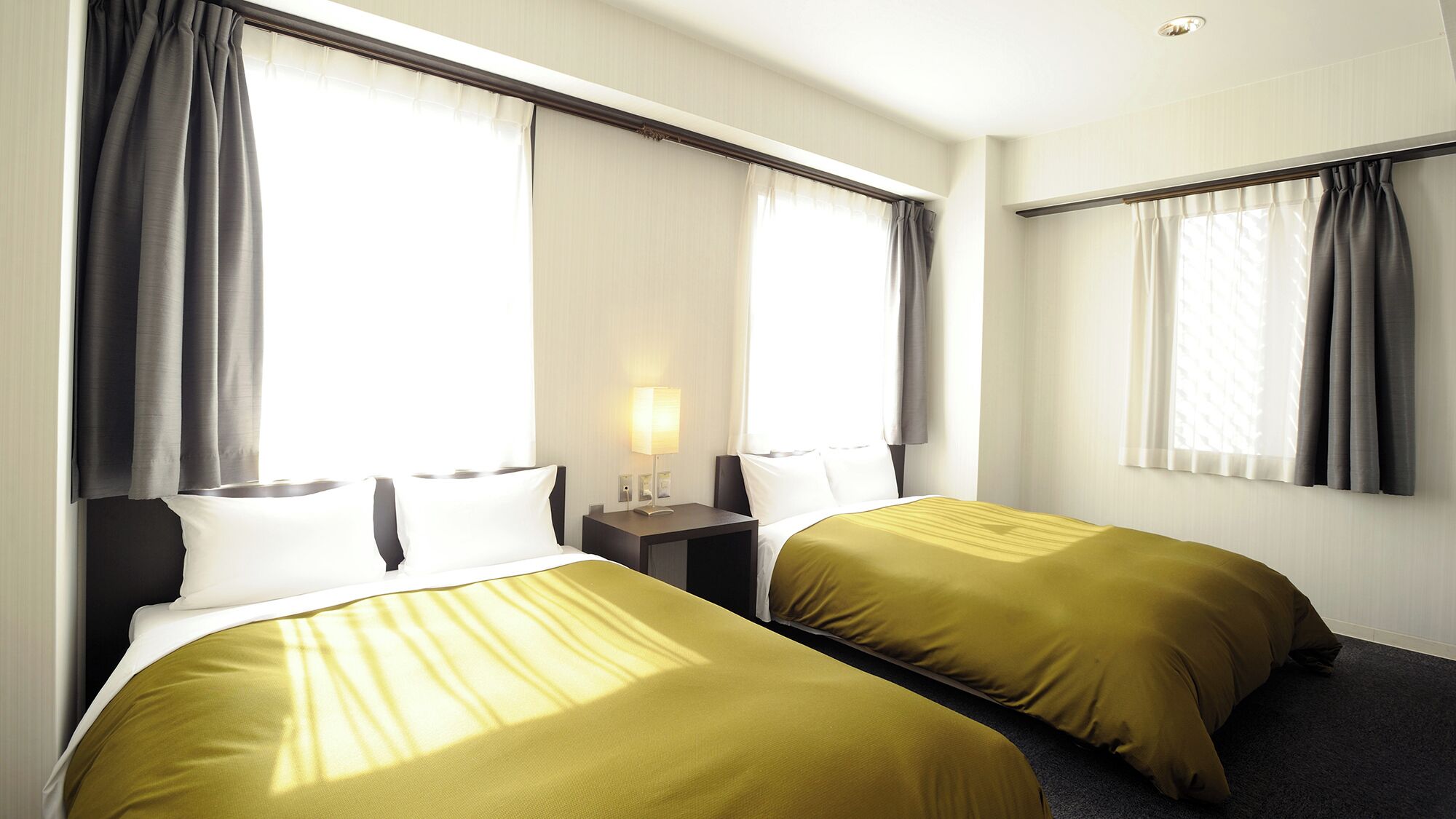 A hotel room with two beds, a nightstand with a lamp, and large windows with curtains.