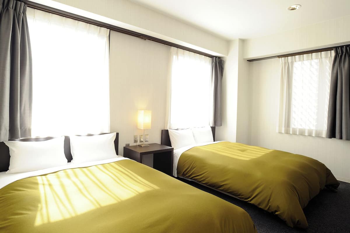 Une chambre d’hôtel avec deux lits, une table de chevet avec une lampe et de grandes fenêtres avec des rideaux.