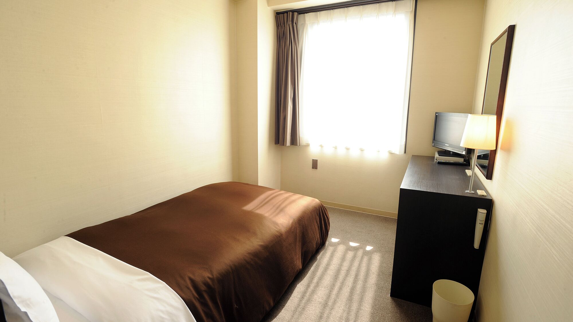 A hotel room with a bed, a desk with a TV, a lamp, and a mirror.