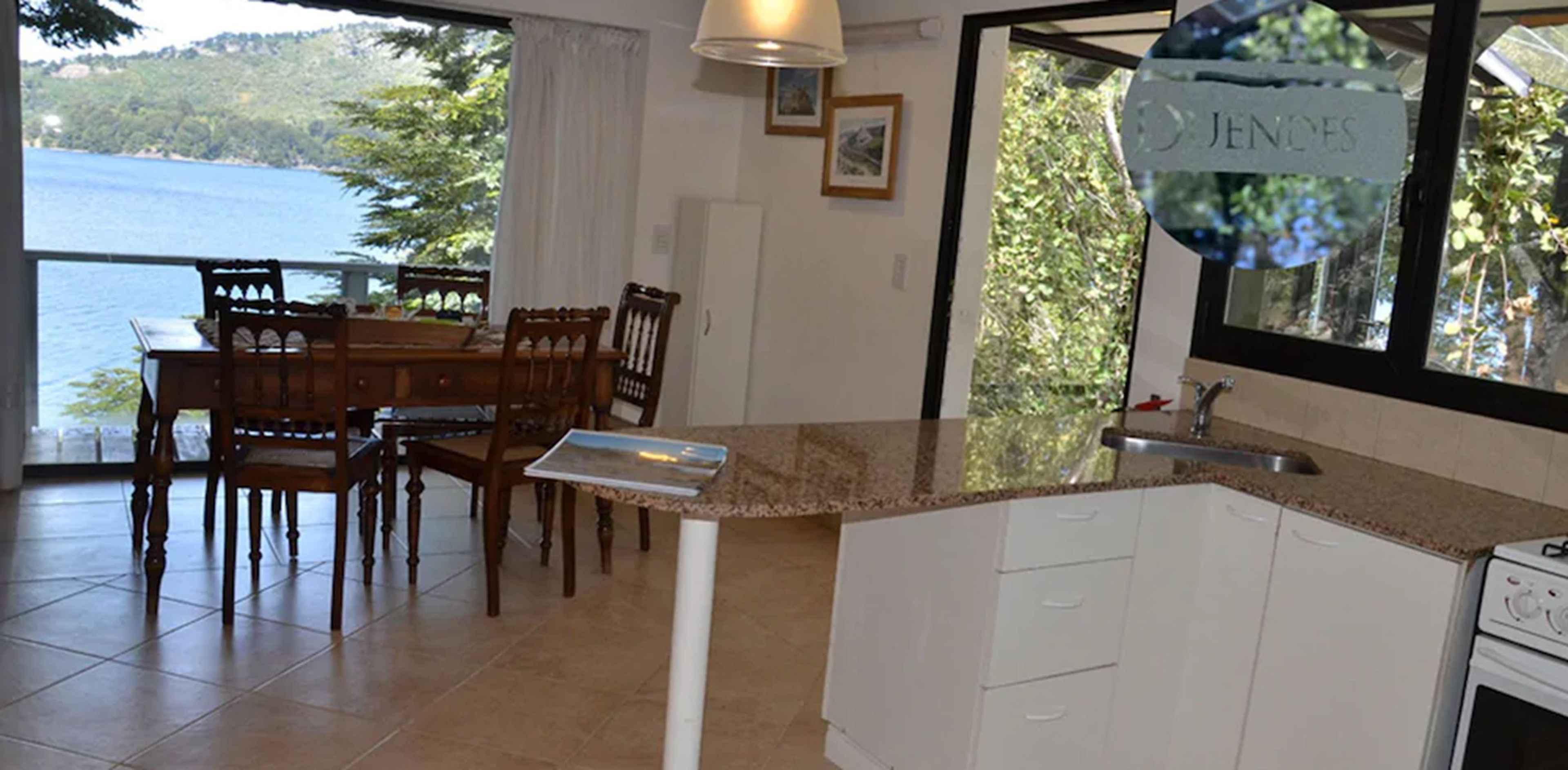 A dining area with a table and chairs, a kitchen with a sink and stove, and a view of a lake through large windows.