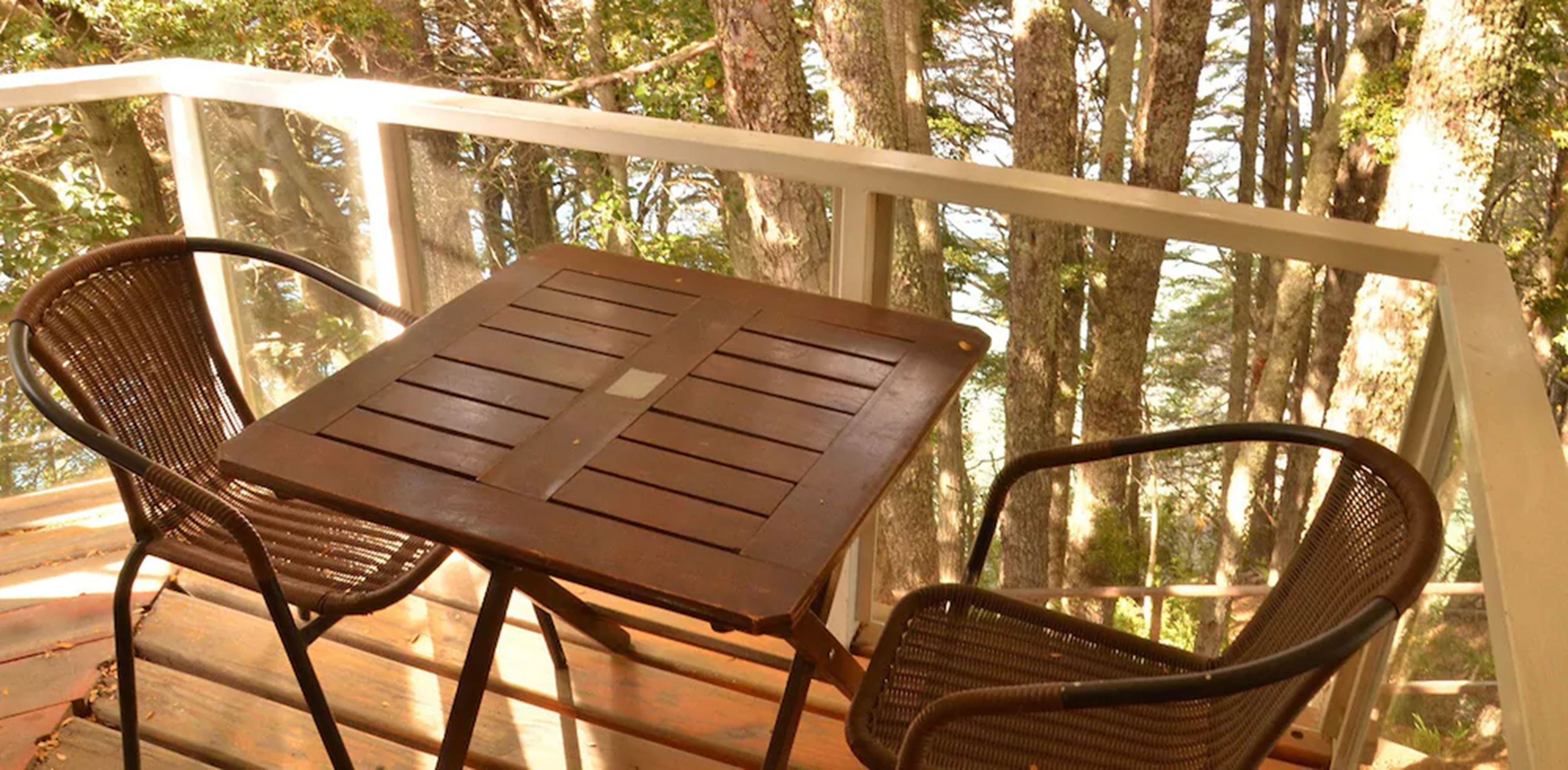 A wooden table and two wicker chairs on a wooden deck with a white railing, surrounded by trees.