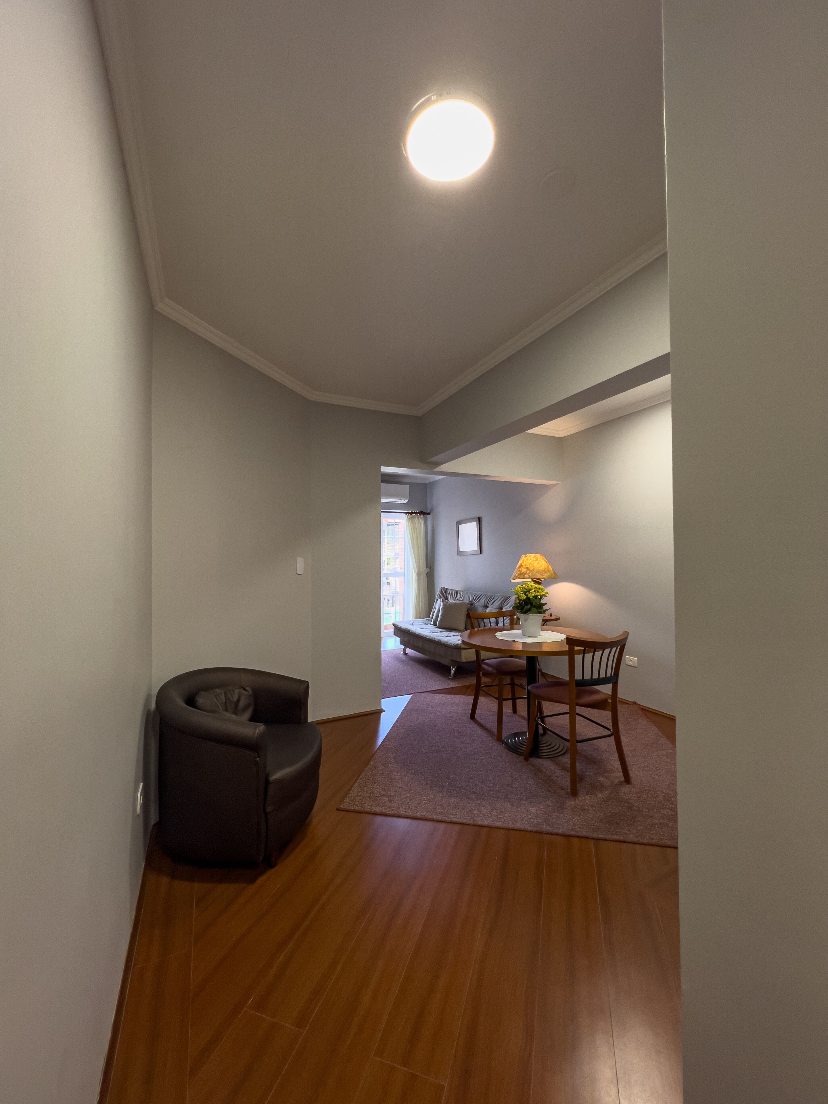 A narrow hallway with a dining area at the end, featuring a table with chairs and a lamp.