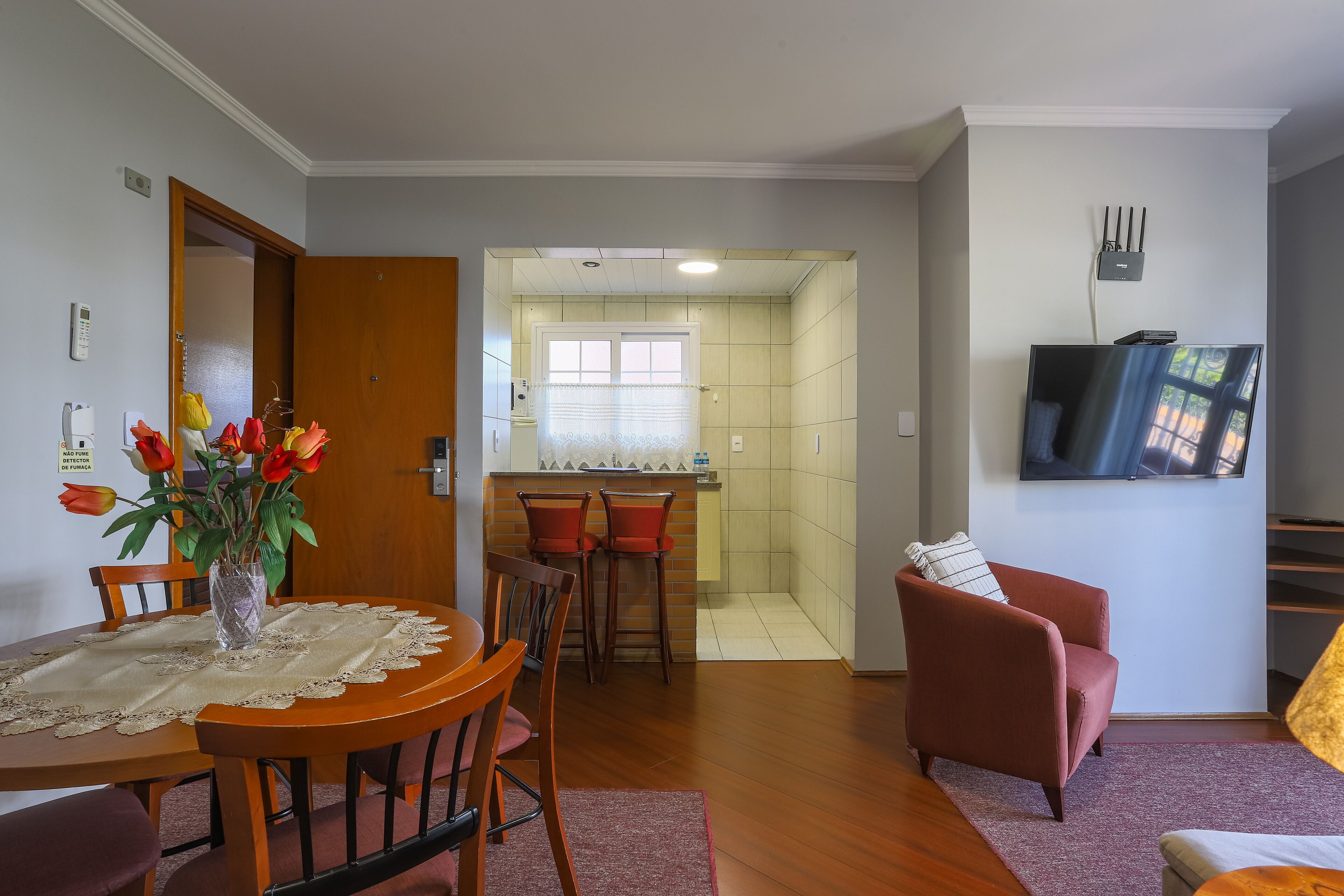 Un comedor con una mesa, sillas y un florero. Se observa una cocina al fondo con una ventana y una puerta.