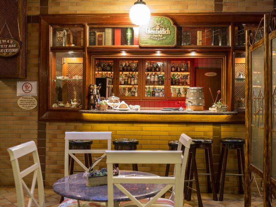 A bar with a brick wall, wooden shelves, and a counter with various bottles and glasses. There are three stools and two chairs around a small table.