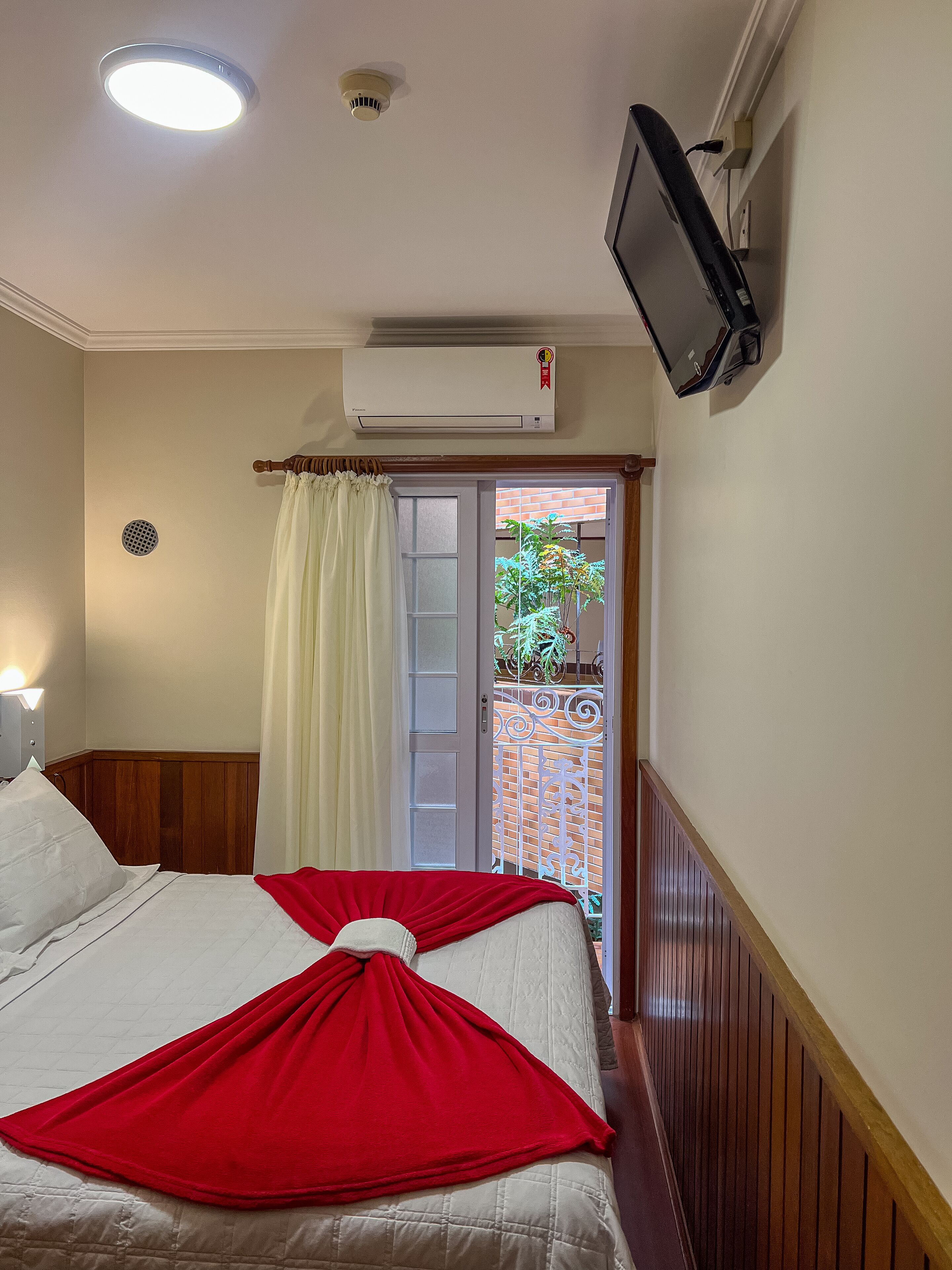 A hotel room with a bed, a TV mounted on the wall, an air conditioning unit, and a window with a curtain.