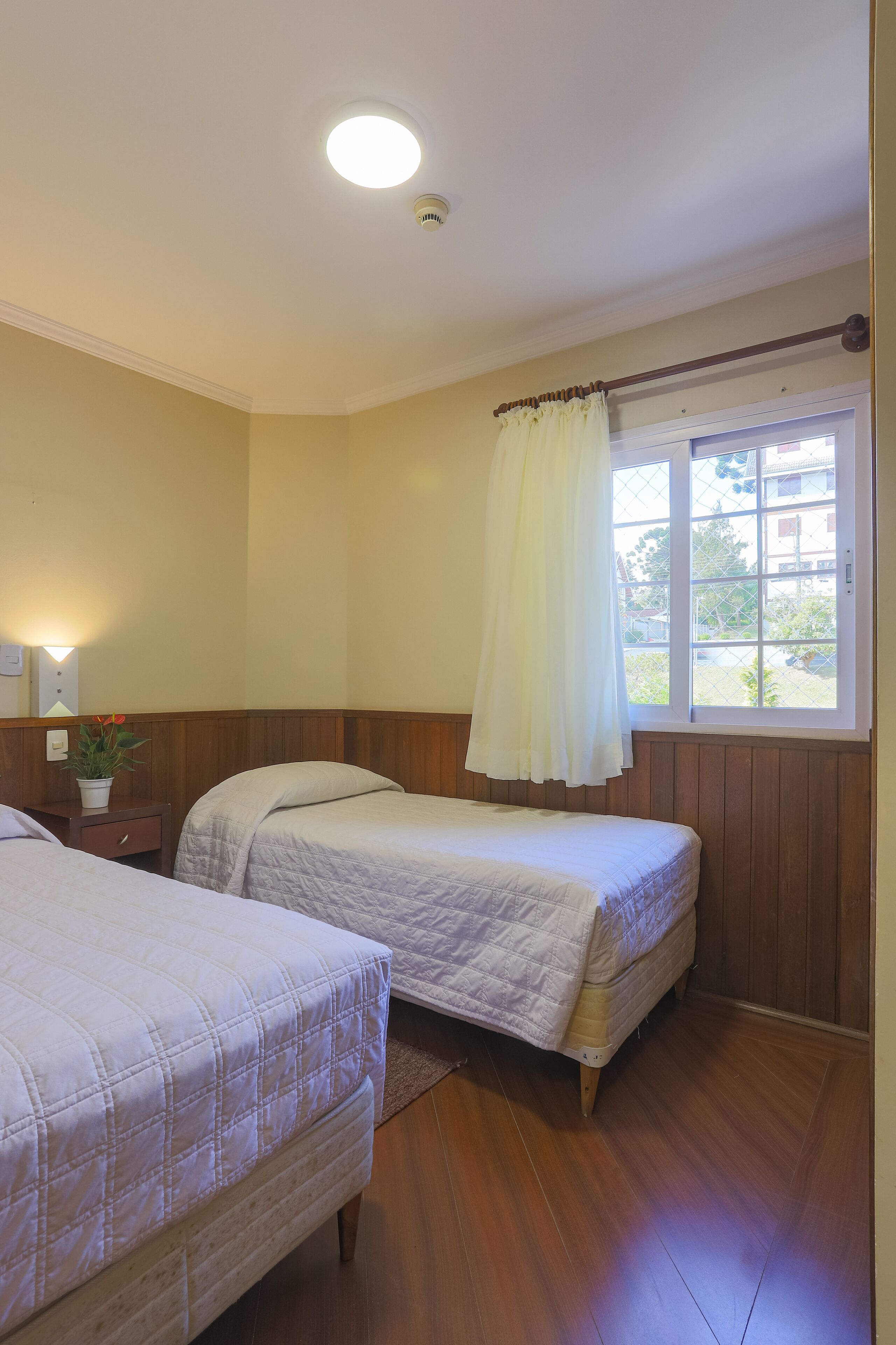 Ein Schlafzimmer mit zwei Betten, Holzboden, einem Fenster mit Vorhang und einer Deckenleuchte.