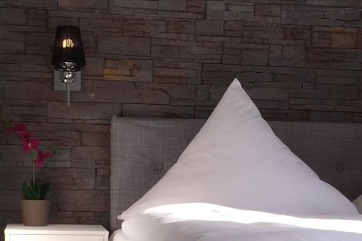 A neatly made bed with white linens, a grey headboard, and a nightstand with a potted plant beside it.