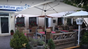 An outdoor seating area with tables and chairs under umbrellas, potted plants, and a building with a sign reading "Achilles Grill". - Hotel Restaurant Achilles (Kirkel)