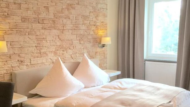 A hotel room with a brick-patterned wall, a bed with white bedding, two bedside lamps, and a window with curtains. - Hotel Restaurant Achilles (Kirkel)