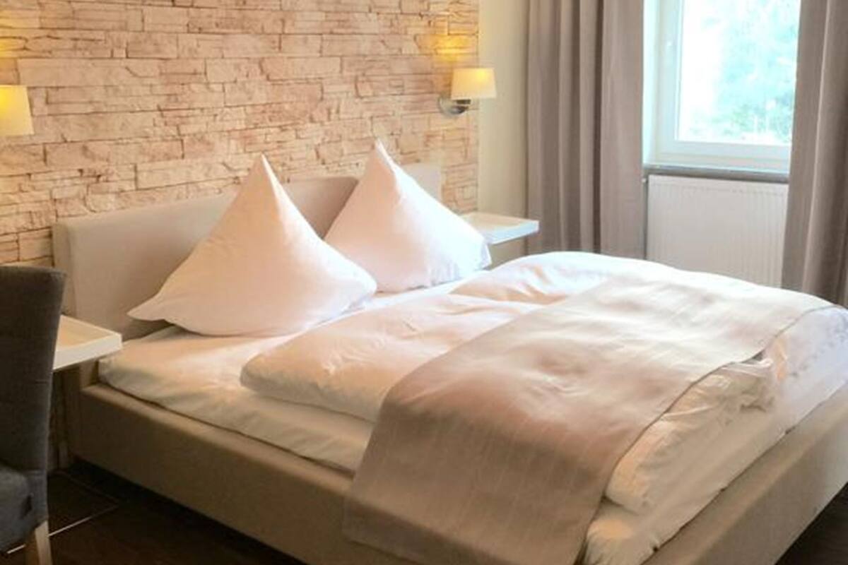 A hotel room with a brick-patterned wall, a bed with white bedding, two bedside lamps, and a window with curtains.