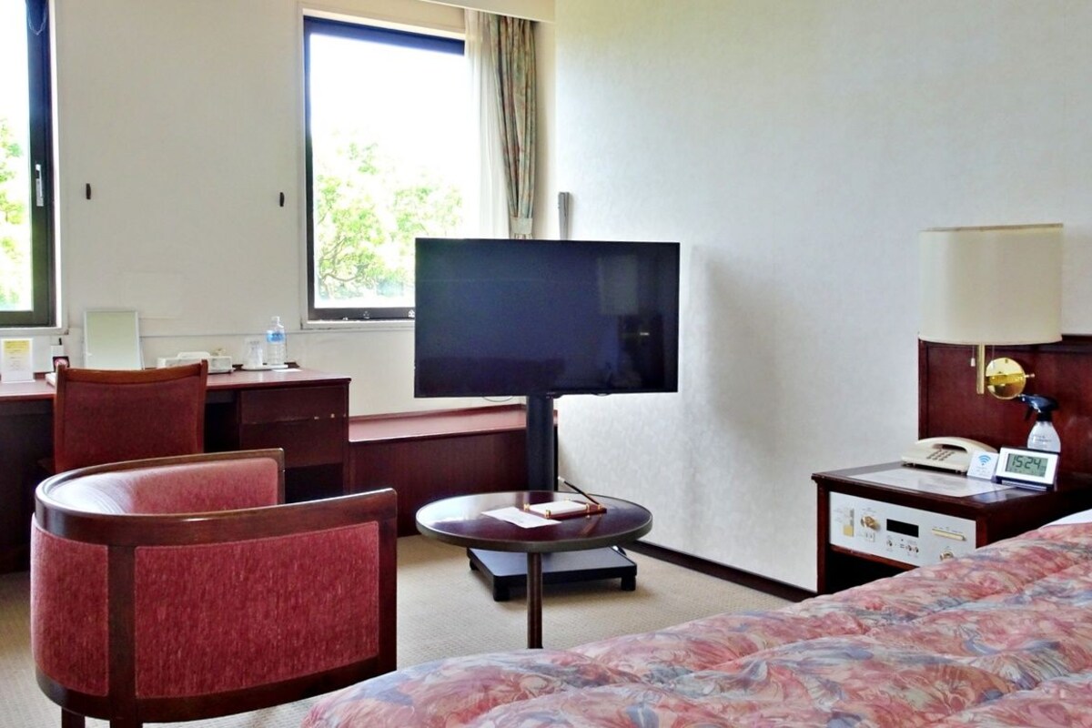 A hotel room with a bed, a desk with a chair, a television, a bedside table with a lamp, and a window with a view of trees.