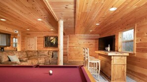 A wooden cabin interior with a pool table, a sofa, a dining area, and a TV.