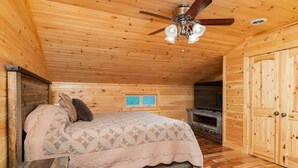 A wooden cabin bedroom with a bed, a ceiling fan, a television, and a wooden wardrobe.