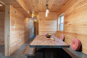 A wooden cabin interior with a dining table, chairs, a bowl of fruit, and a window.