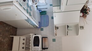 Et kompakt køkken med komfur, mikrobølgeovn og vask. Der er en lille bordplade og en blå stol.