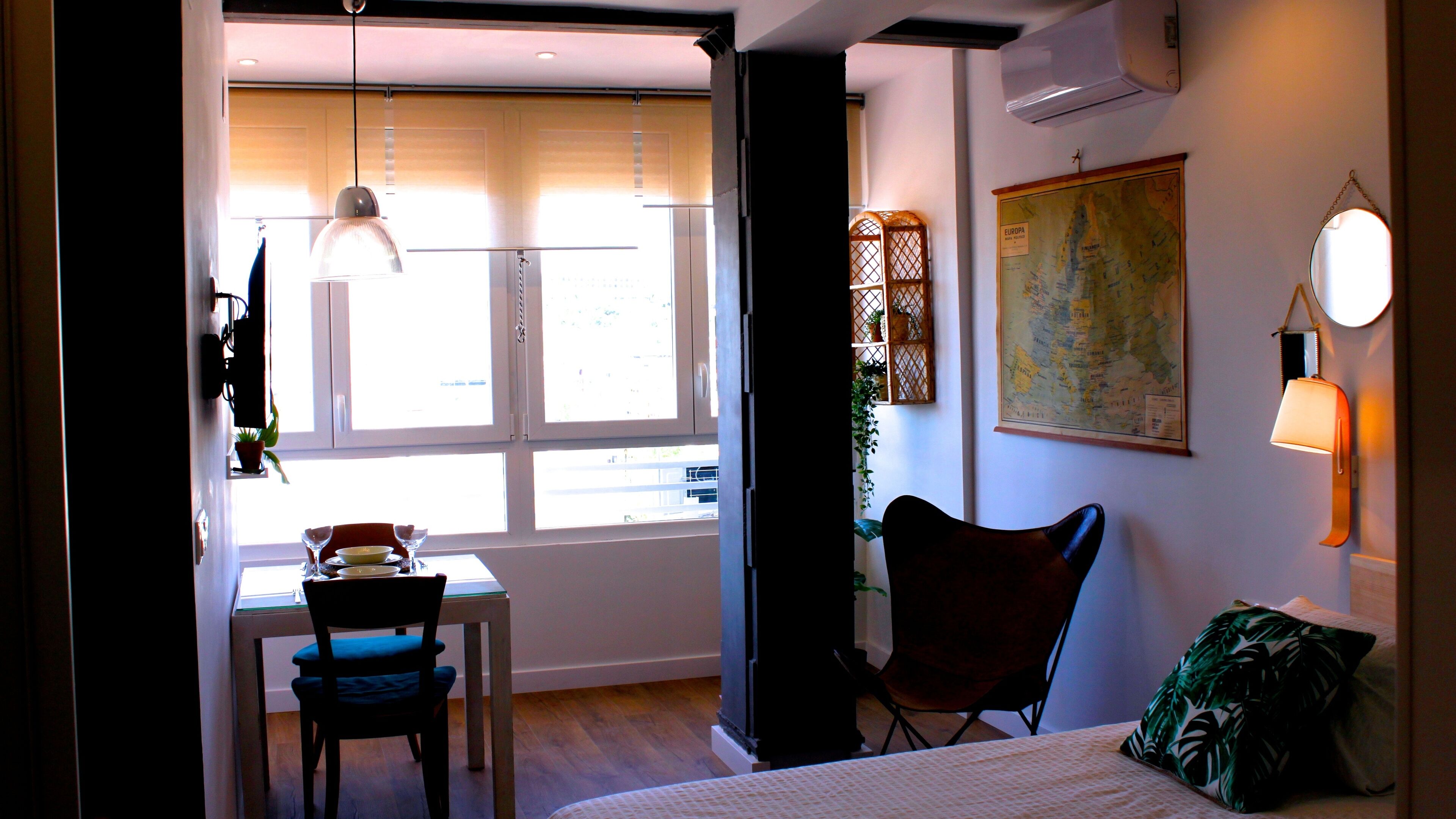 A compact room with a dining table, chairs, a bed, a map on the wall, and a window with blinds.