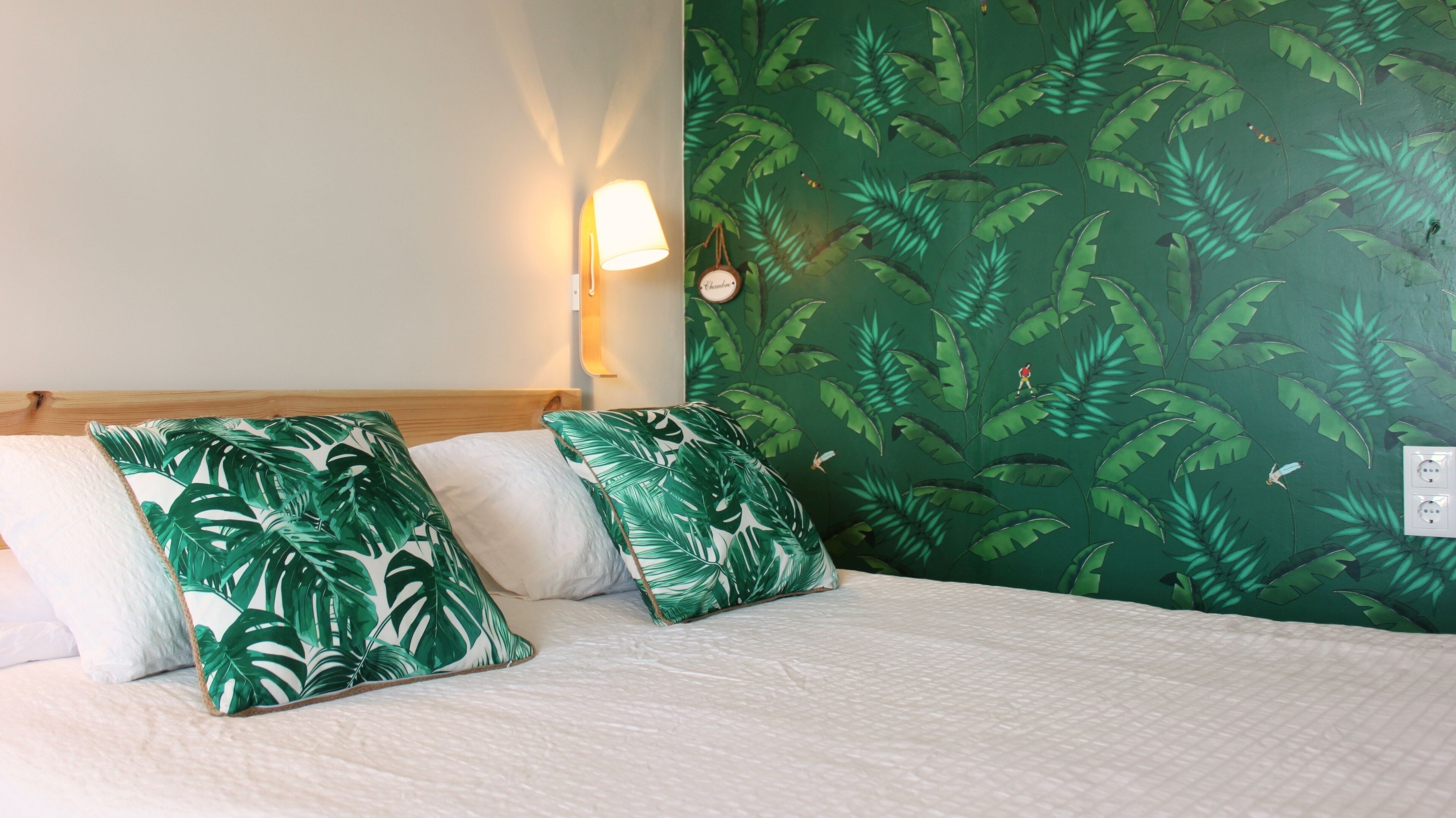 Ein Bett mit weißer Bettwäsche und Kissen mit tropischen Blättern, ein Holz-Kopfteil, eine wandmontierte Lampe und eine Wand mit Blättermotiv.