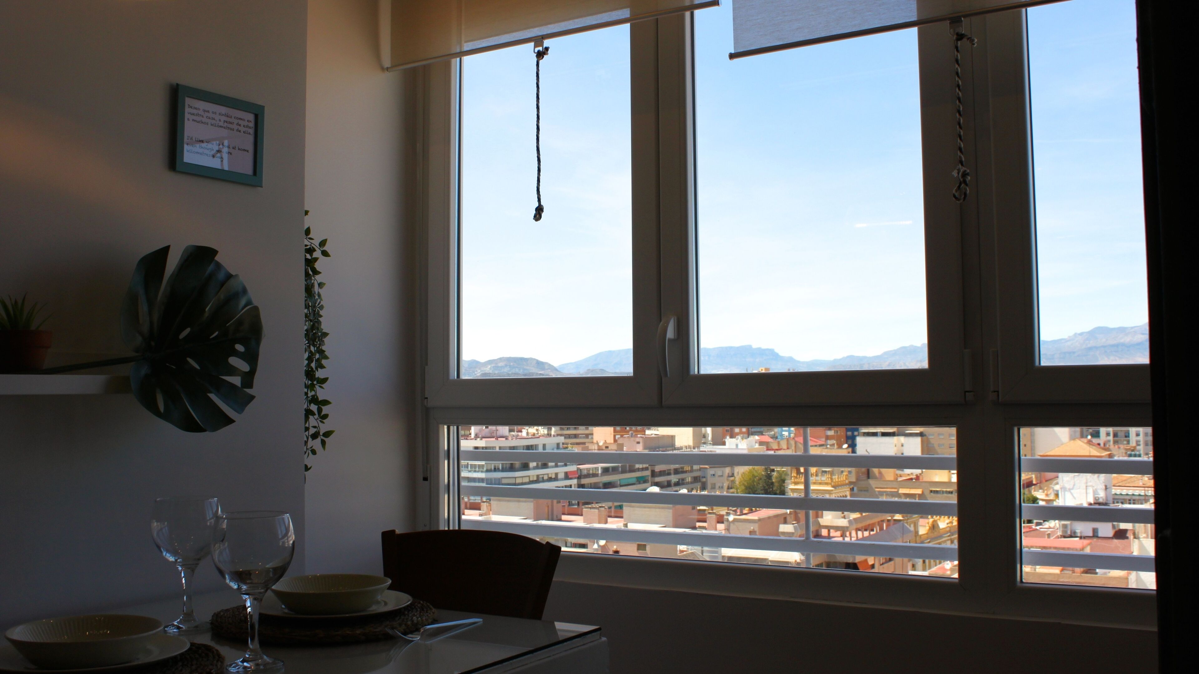 Un comedor con una mesa dispuesta para dos, vista a la ciudad a través de amplias ventanas y una planta en la pared.