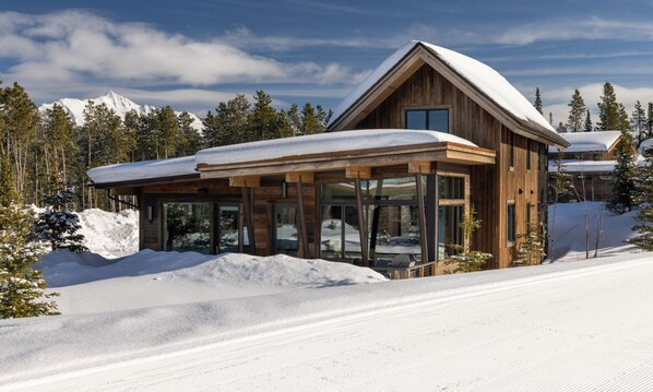 A wooden cabin with a glass extension, surrounded by snow and trees, with mountains in the background.