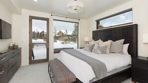 A bedroom with a large bed, bedside tables, a bench, a TV, and a view of a snowy landscape. - Big Sky Vacation Rentals: Silvertip 22 Alpine Bend (big sky)