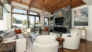 A spacious living room with a stone fireplace, large windows offering a view of a snowy landscape, and a cozy seating area. - Big Sky Vacation Rentals: Silvertip 22 Alpine Bend (big sky)