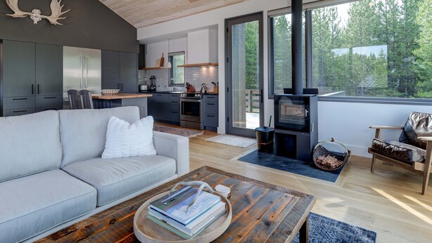 A modern living room with a sofa, coffee table, and books. A kitchen with a stove and cabinets is visible through an open door.