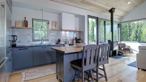 A modern kitchen with grey cabinets, a wooden island, and a large window offering a view of the forest.