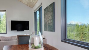 A modern living room with a wooden table, a glass-top coffee table with a candle, a flat-screen TV, and large windows offering a mountain view.