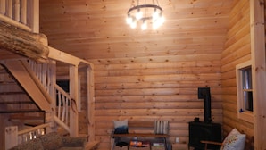A log cabin interior with a wooden staircase, a sofa, a chair, a table, and a fireplace. - Jackson, NH Log Cabin near Storyland in White Mountains: 2Queen Bdrms+Sleep Loft (Jackson)