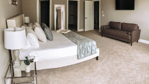 A bedroom with a large bed, a bedside table with a lamp, a sofa, and a TV on the wall.