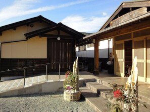 Il s'agit d'un bâtiment japonais traditionnel, avec une façade en bois, un chemin de pierre menant à l'entrée et un petit jardin agrémenté d'une lanterne en pierre et de fleurs.