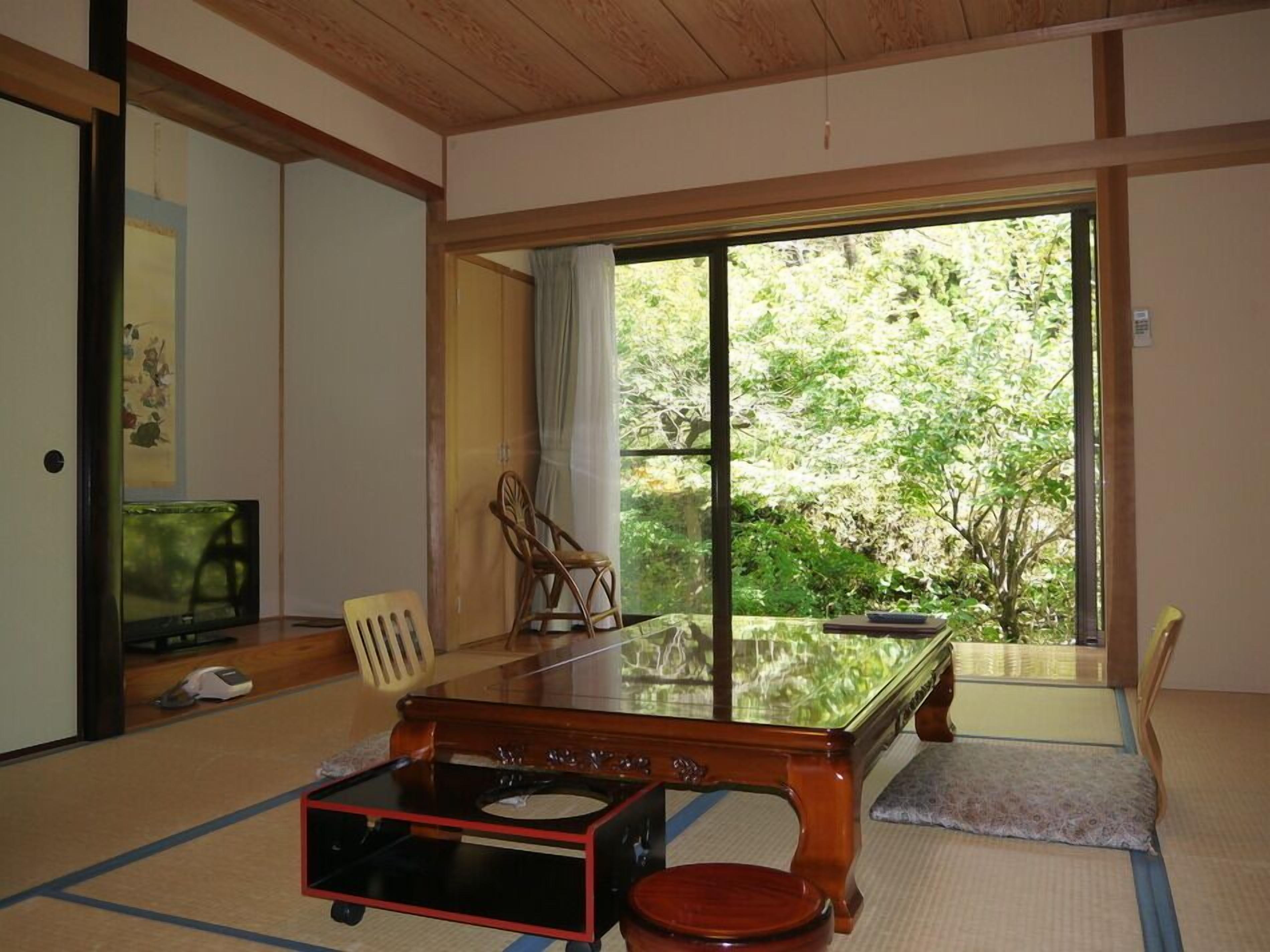 A traditional Japanese room with tatami flooring, a low wooden table, a television, and a sliding glass door leading to a garden.