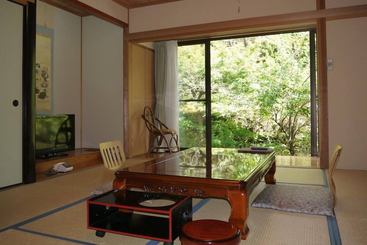 Ein traditioneller japanischer Raum mit Tatami-Bodenbelag, einem niedrigen Holztisch, einem Fernseher und einer Schiebeglastür, die zu einem Garten führt.