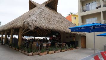 A thatched-roof outdoor dining area with wooden chairs and tables, surrounded by potted plants.