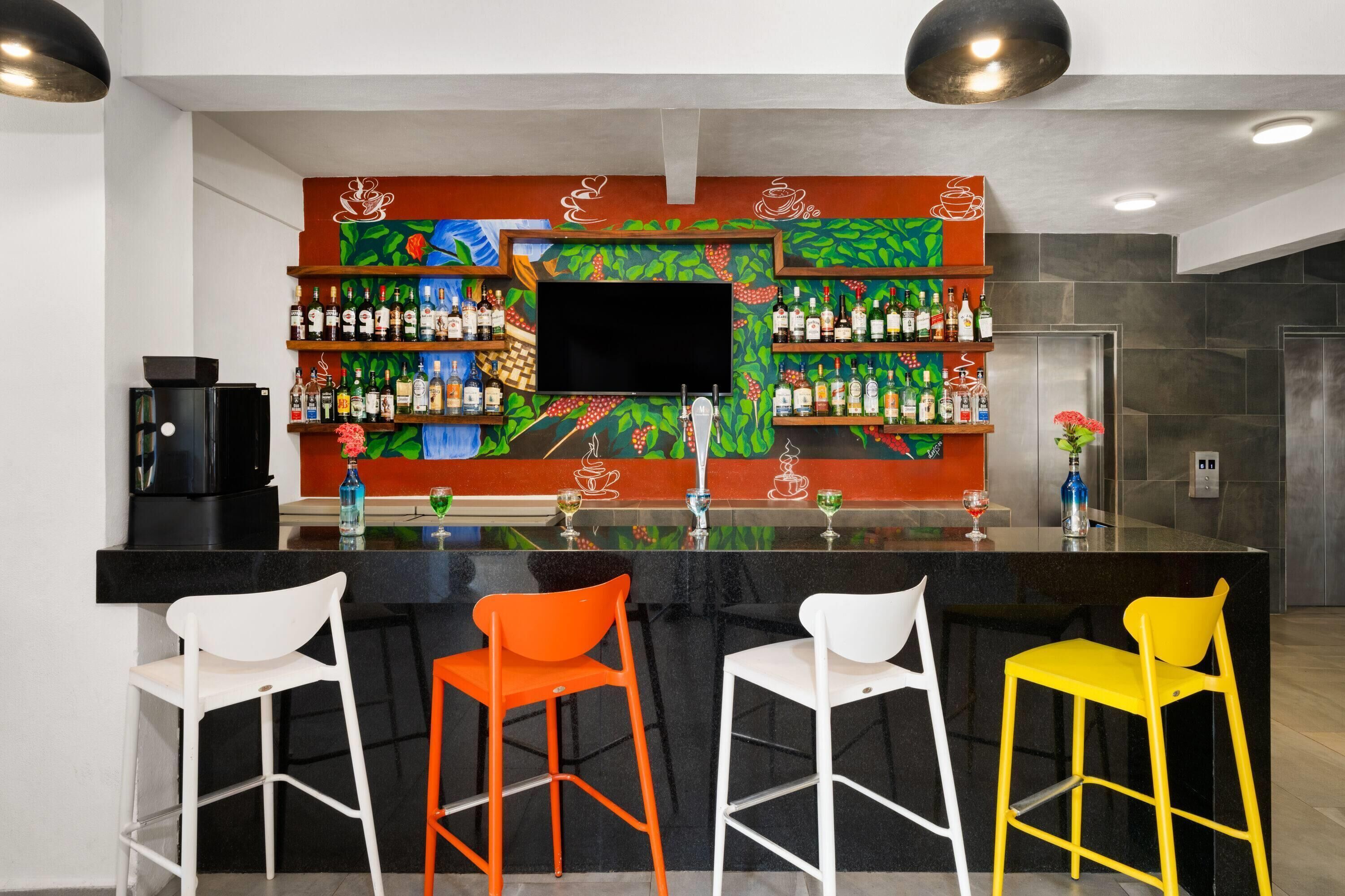 Zona de bar con una televisión de pantalla plana, estantes con botellas de licores y tres taburetes en blanco, naranja y amarillo.