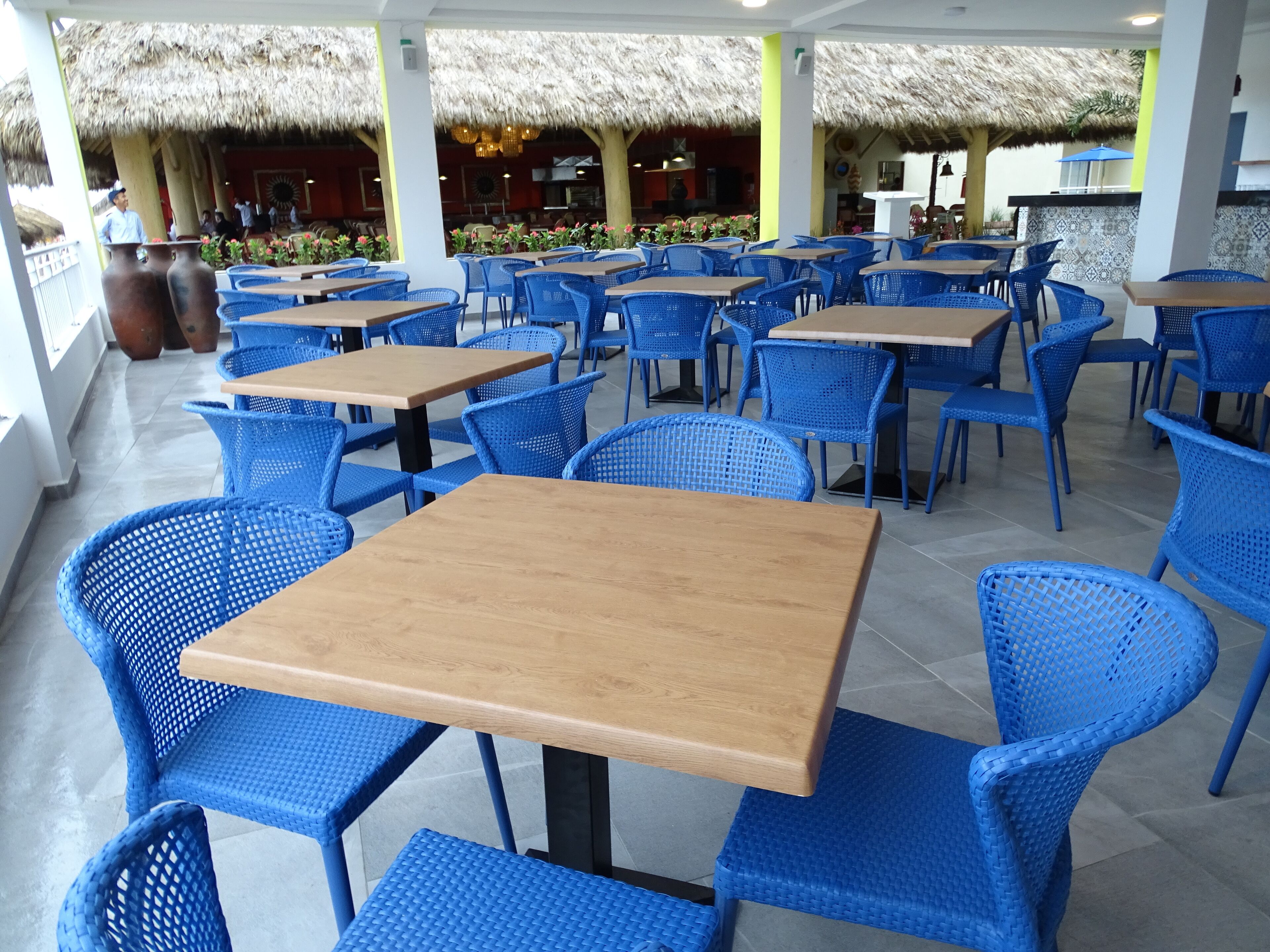 A restaurant with blue chairs and wooden tables, thatched roof, and potted plants.