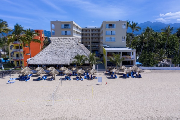 Un resort en la playa con una construcción de techo de paja, sillas de descanso azules y palmeras.