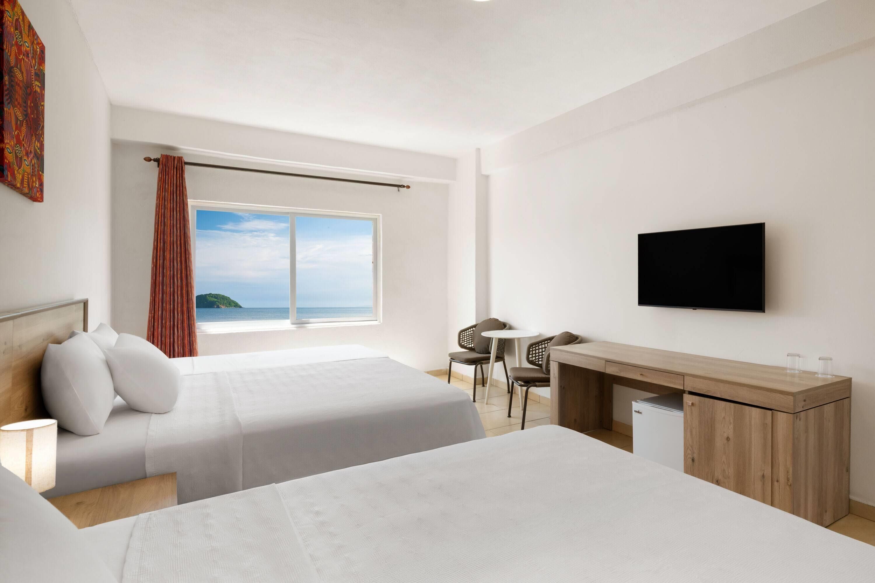 A hotel room with two beds, a desk, a TV, and a window with a view of the sea.