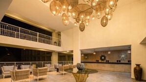 A spacious lobby with a high ceiling, a central glass table, wicker chairs, and a decorative chandelier.