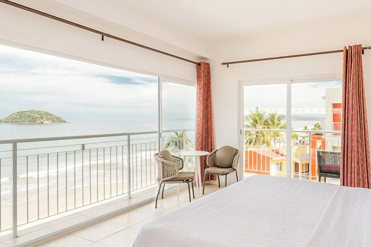 A hotel room with a large bed, a balcony with a view of the sea, and a small table with chairs.
