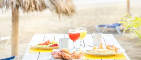 Mesa en la playa con comida y bebidas, dos sombrillas de paja y vistas al océano.