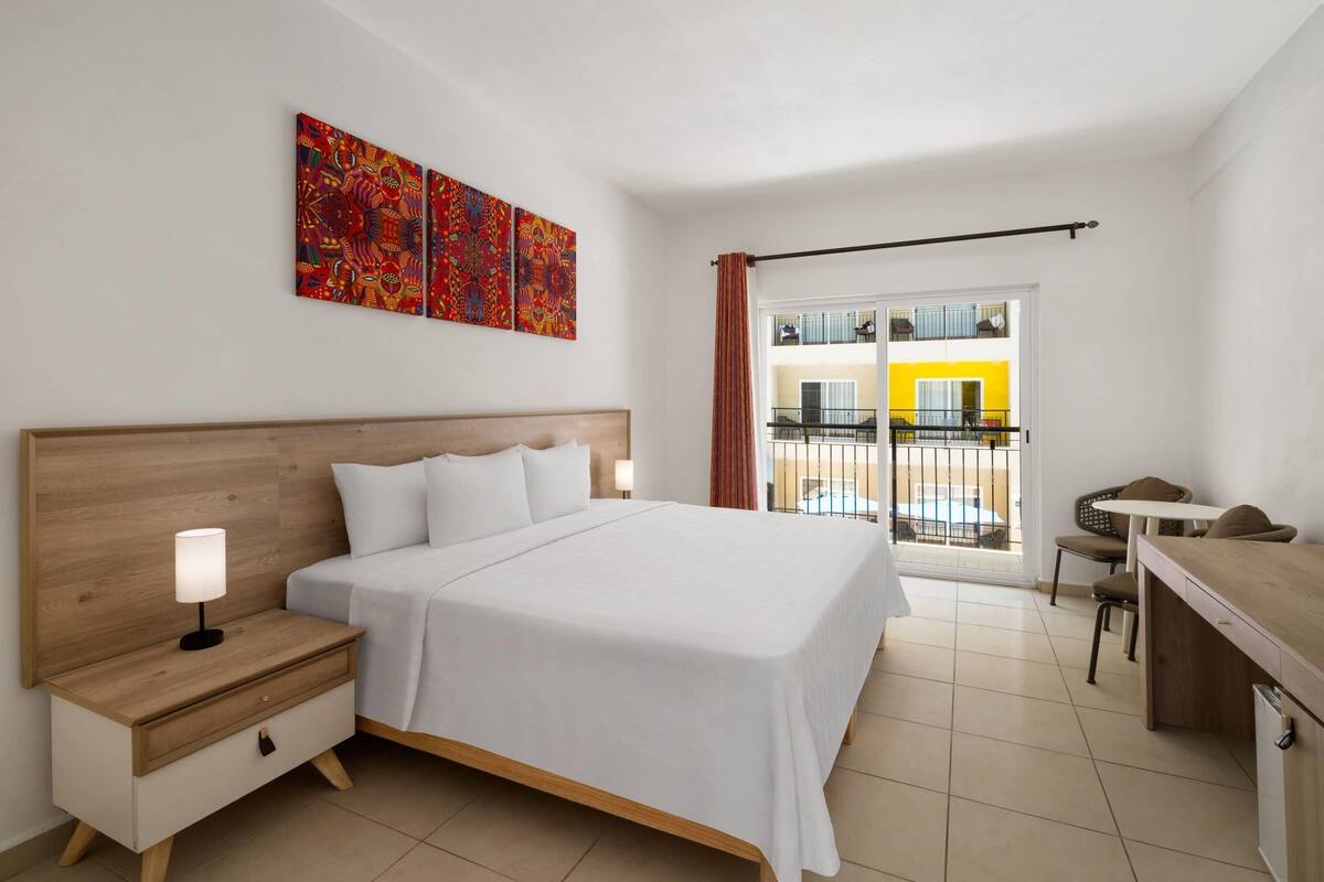 A hotel room with a bed, bedside table, lamp, artwork, and a view of a balcony with a yellow building.
