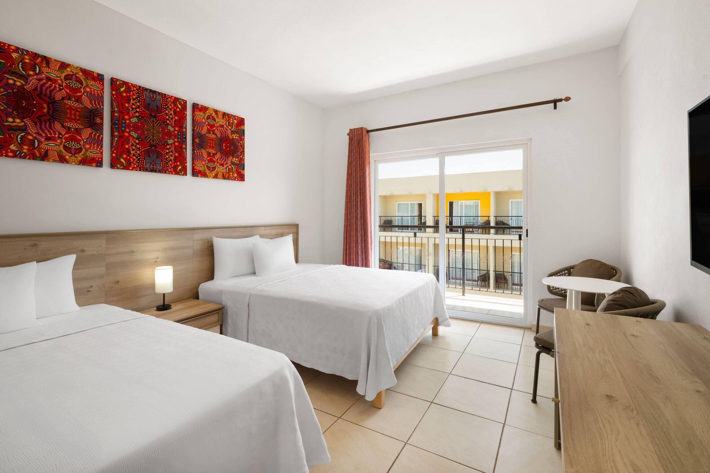Habitación de hotel con dos camas, cabecera de madera, escritorio, silla, televisor y balcón con vistas a edificios.