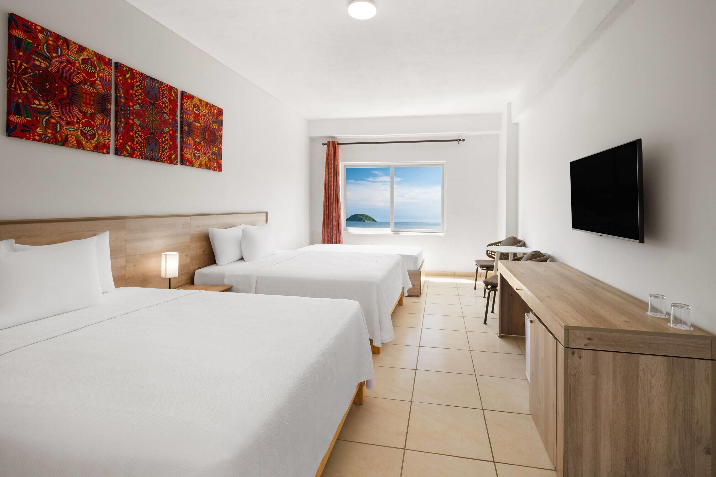 Habitación de hotel con dos camas, un escritorio, un televisor y una ventana con vista al mar.