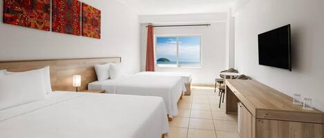 A hotel room with two beds, a desk, a TV, and a window with a view of the sea.