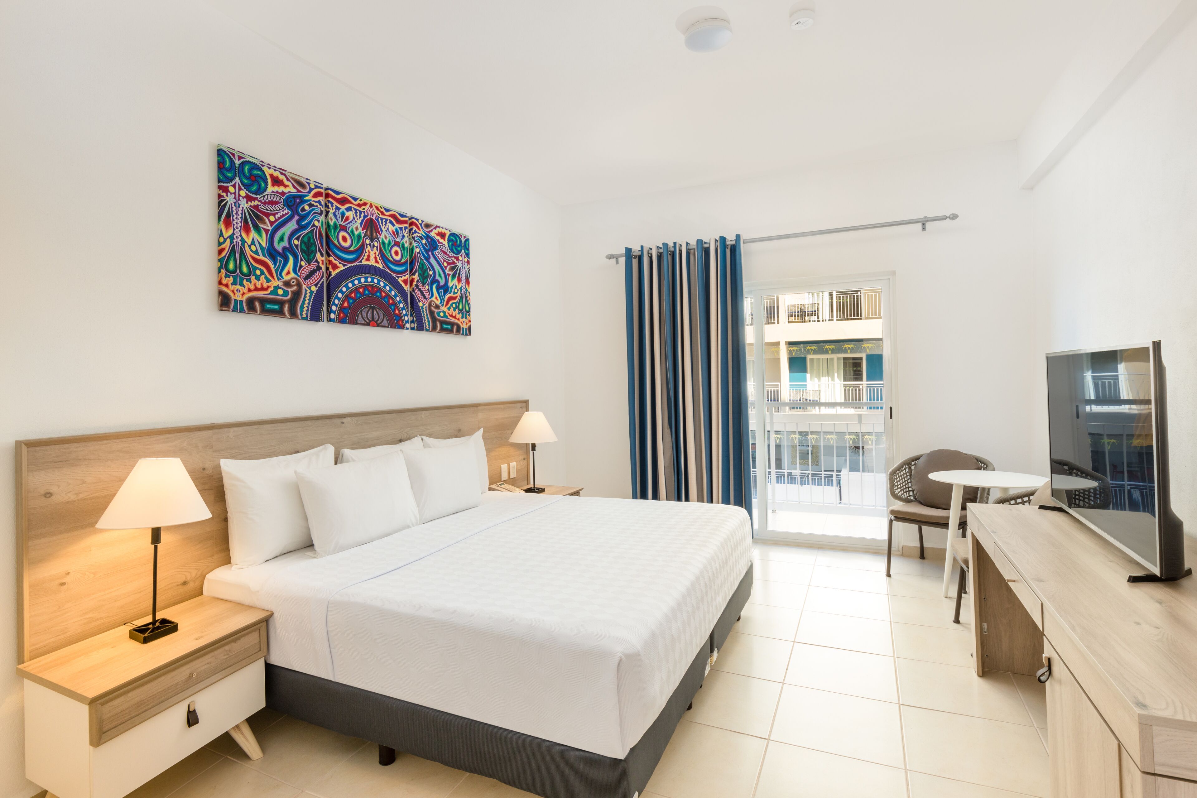 Quarto de hotel com cama grande, escrivaninha com televisão, mesa pequena e varanda com cortinas.