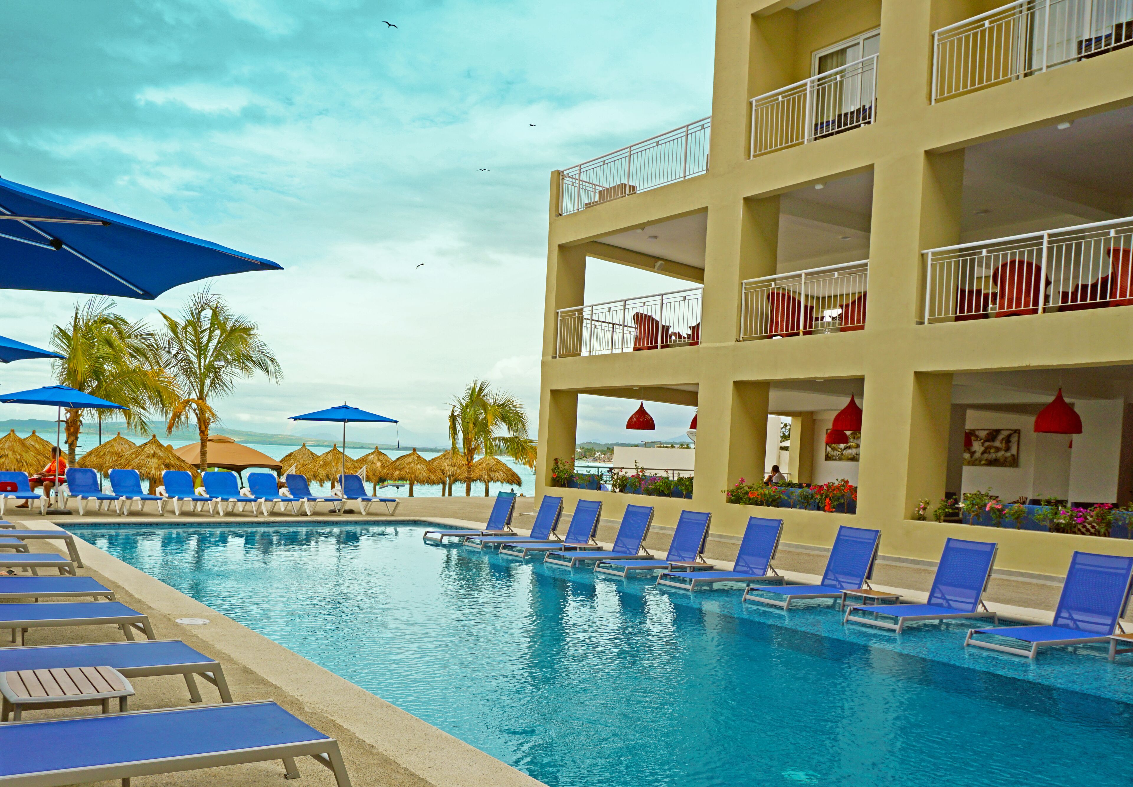 Área de piscina com espreguiçadeiras e guarda-sóis, um edifício com várias andares e sacadas, e céu claro.