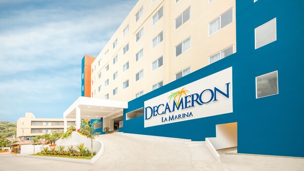Un hotel llamado Decaméron La Marina con un diseño moderno, que presenta una fachada azul y beige destacada, un acceso cubierto y un área ajardinada con plantas.