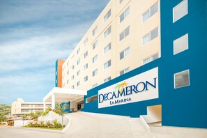 Ein Hotel namens Decaméron La Marina mit modernem Design, das eine markante blaue und beige Fassade, einen überdachten Eingangsbereich und eine angelegte Grünfläche mit Pflanzen zeigt.