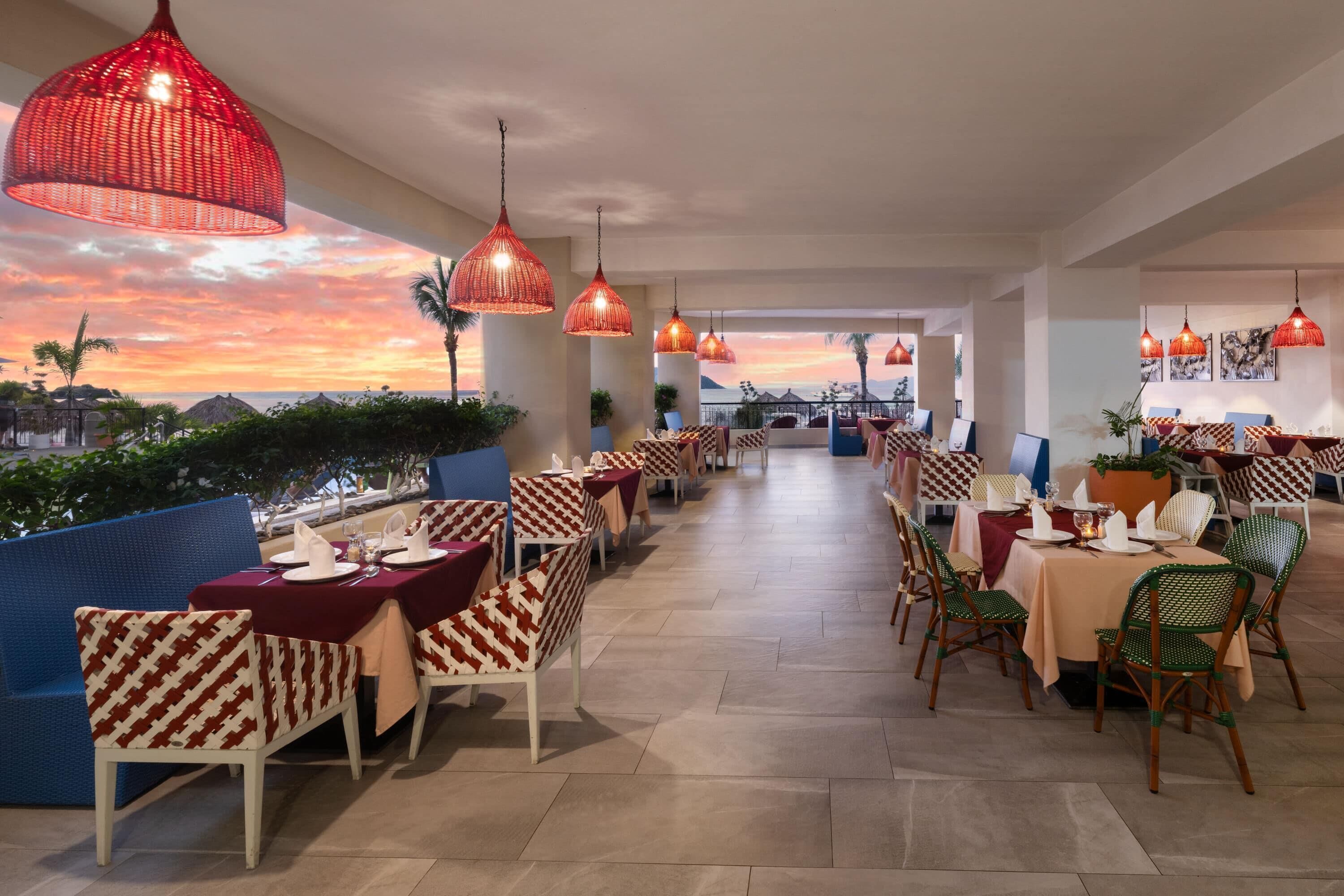 Un restaurante con mesas dispuestas para comer, luces colgantes rojas y vistas al atardecer.